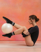 Woman in black outfit with black striped yoga pilates grip socks holding a white ball against an orange background